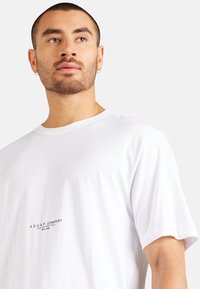 Weißes Baumwoll-T-Shirt mit Rundhalsausschnitt und kleinem schwarzem Grafiklogo auf der Brust. Kurze Ärmel; glatte, leichte Textur.