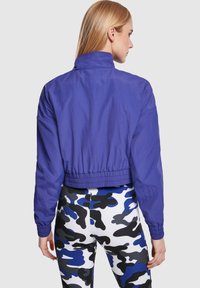 Kuttet lilla vindjakke med høy krage, elastisk liv, og lange ermer, kombinert med svarte, hvite og blå camo leggings.