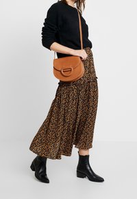 Zwarte trui, enkellange rok met bloemenpatroon in donkere tinten, bruine leren schoudertas met een cirkelvormig detail, en zwarte enkellaarsjes.