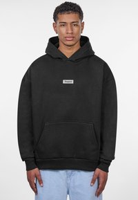 Haut à capuche noir en tissu doux avec une poche avant, présentant un petit patch rectangulaire au centre et des poignets côtelés.