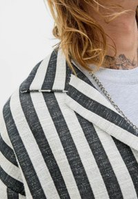 Veste rayée en tissu tissé noir et blanc, avec une finition texturée et un col cranté. Détails en chaîne argentée sont visibles.