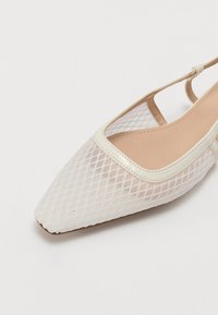 Valkoinen verkko slingback-kenkä, jossa on terävä kärki, kermaiset yksityiskohdat ja teksturoitu timanttikuvio. Varustettu hoikalla nilkkaremmillä ja beigellä sisäosalla.