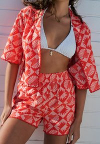 Completo corto a motivo rosso con camicia oversize abbinata, caratterizzato da un design a scallop e bikini bianco texturizzato sottostante.
