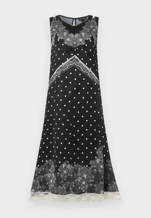 Žena s tmavými vlasmi v čiernych midi šatách bez rukávov s bielymi bodkami, kvetinovým čipkovým vzorom a držiaca malú čiernu kabelku.