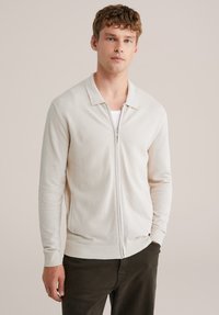 Cardigan na zips v svetlo béžovej farbe, s textúrovaným pleteným vzorom, golierom, rebrovanými manžetami a dvoma bočnými vreckami. Nosený cez biele tričko a tmavé nohavice.