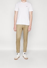T-shirt en coton blanc avec un logo noir "HUGO", associé à un pantalon slim beige clair et des baskets blanches avec une semelle en caoutchouc.