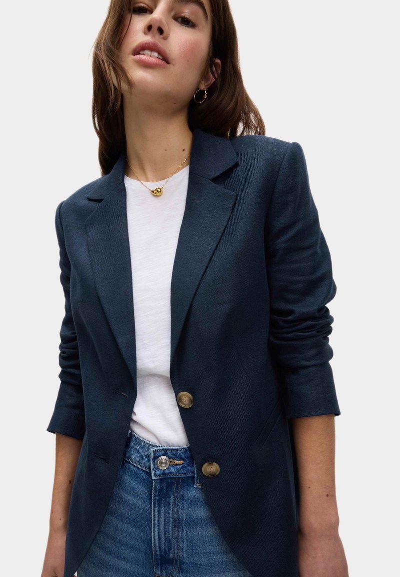 Blazer bleu marine avec deux boutons en laiton, coupe ajustée, texture douce, associé à un t-shirt blanc et un jean en denim.