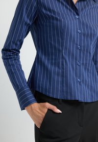 Camicia blu con bottoni, a righe bianche verticali, maniche lunghe e taglio adattato. Tessuto liscio e colletto a punta con maniche con polsini.