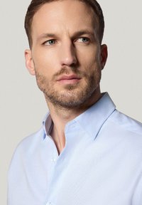 Jasnoniebieska koszula z kołnierzykiem o strukturalnym kształcie, z delikatną fakturą i subtelnym połyskiem, uzupełniona przez przezroczyste guziki.