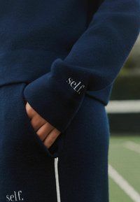 Vêtement en polaire bleu marine à texture brossée, avec une poche et une ligne d'accent blanche. Logo « self. » brodé en argent sur le poignet.