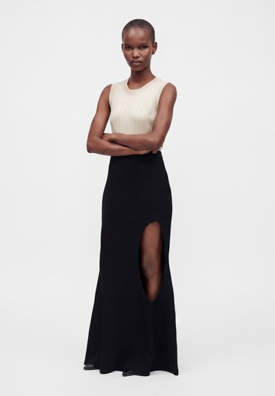 Woman stands with arms crossed wearing a sleeveless cream top and long black skirt with a high slit on one leg against plain background.