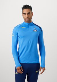 Langærmet atletisk top i livlig blå med halv lynlås i nakken. Har et mørkeblåt skulderpanel og et klublogo på brystet.