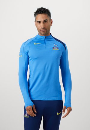 Langærmet atletisk top i livlig blå med halv lynlås i nakken. Har et mørkeblåt skulderpanel og et klublogo på brystet.