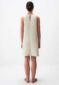 Ärmelloses, hellbeiges Kleid mit einem Rückenschlitz, glatter Textur und knielangem Saum. Es werden flache Sandalen getragen.
