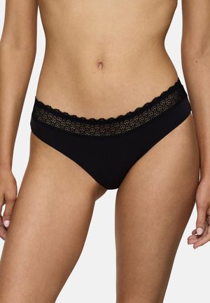 Frau trägt schwarzen Bikini-Unterwäsche mit Spitzenbesatz, zeigt Bauchbereich und obere Oberschenkel, Hände locker an den Seiten.