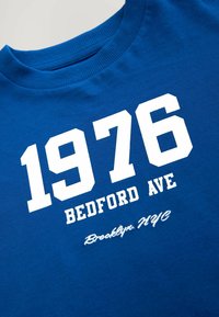 Plava pamučna majica s bijelim blokovnim brojevima "1976" i tekstom "BEDFORD AVE". "Brooklyn, NYC" u kurzivnom pisanju ispod. Teksturirani materijal.