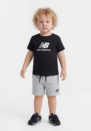 Bambino con capelli biondi ricci che indossa una maglietta nera New Balance, pantaloncini grigi e scarpe da ginnastica nere New Balance, in piedi su uno sfondo bianco.