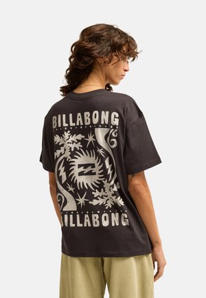 Personne aux cheveux bouclés portant un t-shirt noir Billabong avec un motif graphique blanc et un texte au dos, accompagné d'un pantalon clair.