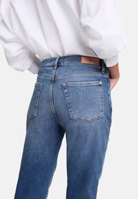 Jeans in denim blu con una finitura sbiadita, cinque tasche e una toppa in pelle marrone sulla vita. Il design è a gamba dritta con un taglio medio.