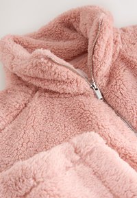 Giacca in pile rosa con una texture morbida e soffice, caratterizzata da un collo alto e un dettaglio con cerniera argentata. Il tessuto è vellutato e accogliente.