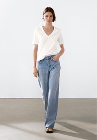 Biała koszulka z dekoltem w serek i krótkim rękawem, zestawiona z jasnoniebieskimi spodniami o szerokich nogawkach. Model stoi na neutralnym tle, z rękami w kieszeniach.