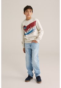 Krämfärgad sweatshirt med röd, blå och vit grafisk design, tillsammans med ljusblå jeans och marinblå skor. En enkel, avslappnad outfit.