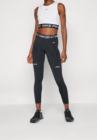 Czarne legginsy z wysokim pasem z logo "NIKE PRO" w białym kolorze. W zestawie z białym, bezrękawnym topem typu crop pokazującym logo Nike.