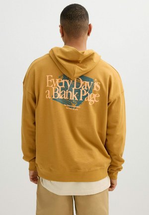 Persona con sudadera con capucha mostaza con capucha bajada, texto "Every Day is a Blank Page" y gráfico de máquina de escribir en la parte trasera, de pie contra un fondo liso.