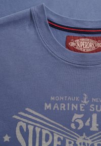 T-shirt bleu à col rond avec étiquette du logo "Superdry" à l’intérieur du col et texte graphique beige délavé "Montauk New Marine Surf 54" sur le devant.