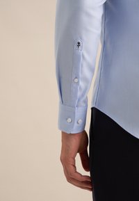 Manica di camicia azzurra chiaro con polsino abbottonato e piccolo ricamo di fiore nero vicino al polso, indossata sopra pantaloni neri.