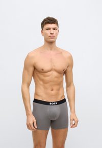 Γκρι μποξεράκια με μαύρη ελαστική μέση που φέρει το λογότυπο "BOSS." Λείο ύφασμα και εφαρμοστό σχέδιο, το μοντέλο εμφανίζεται όρθιο.