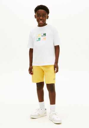 Dreng står smilende, iført hvid grafisk t-shirt, gule shorts, hvide sokker og hvide sneakers på en ensfarvet hvid baggrund.