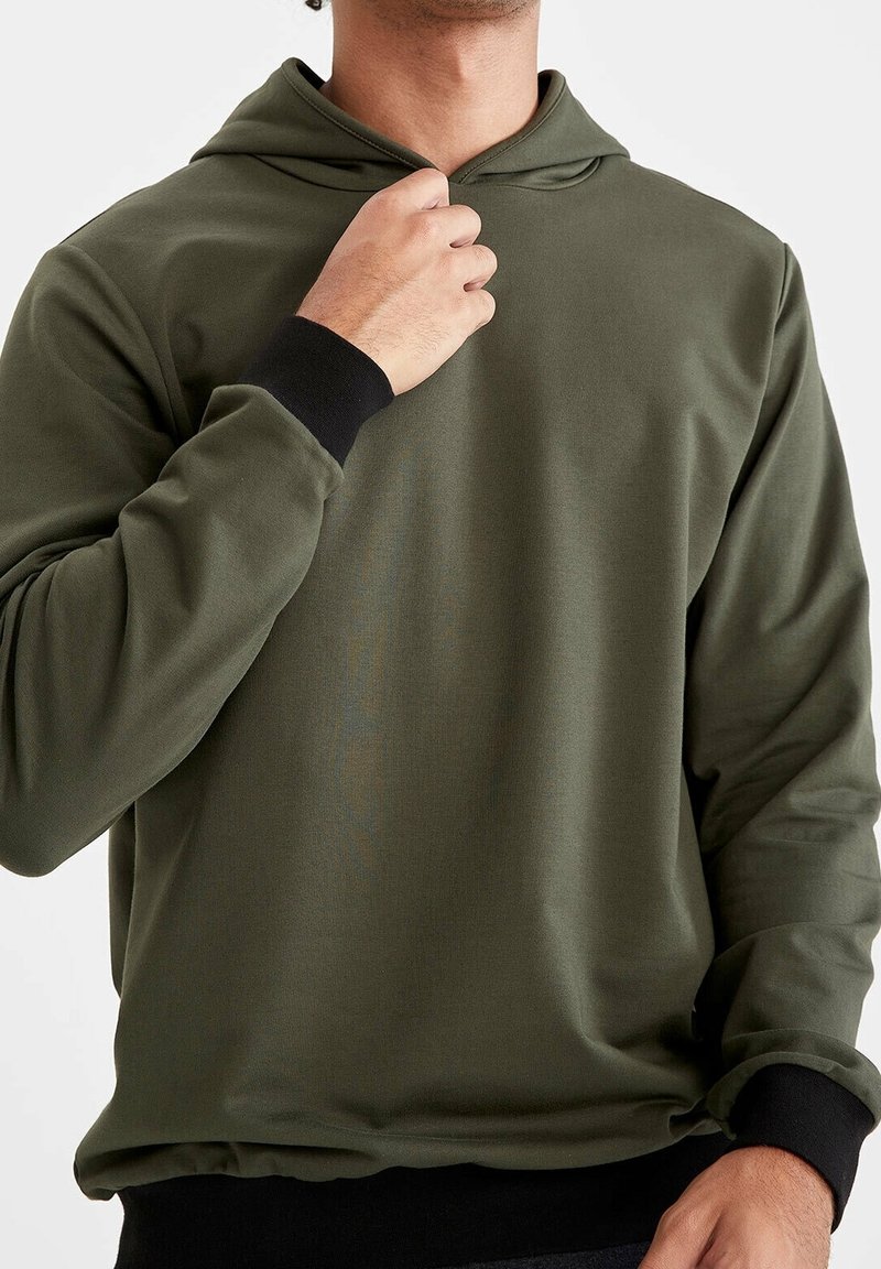 Homme portant un sweat à capuche vert olive avec poignets et ourlet noirs, tenant le col près du cou, sur un fond clair uni.
