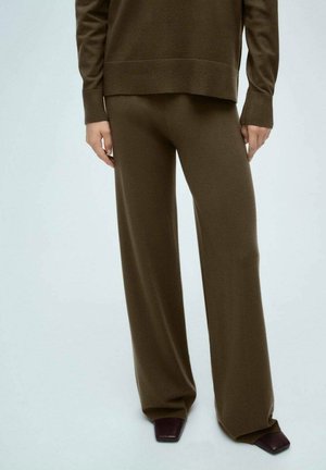 Personne portant un pantalon marron à jambes larges et un haut à manches longues assorti, debout avec les mains détendues le long du corps sur un fond clair.