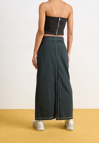 Top noir court sans bretelles avec une fermeture éclair. Jupe maxi rayée vert foncé avec une fente dans le dos et des détails de couture visibles. Chaussures grises chunky.