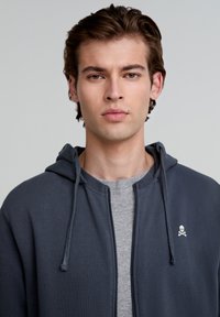 Šedá mikina na zip s kapucí a šňůrkami, s malým bílým logem lebky na levé hrudi, nošena přes světle šedé triko.