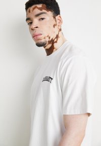 Fehér, rövid ujjú póló, bal oldalon fekete "Carhartt" logóval. Az anyag simának tűnik, szabványos fazonnal.