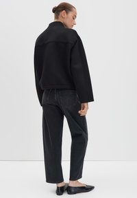 Zwarte cropped jacket met een hoge kraag, gecombineerd met donkergrijze relaxte jeans. Model draagt platte zwarte schoenen. Glad textuur van de stof.