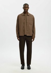 Jeune homme debout, portant une chemise marron à boutons sur un T-shirt assorti, un pantalon marron foncé et des chaussures habillées noires, sur un fond uni.