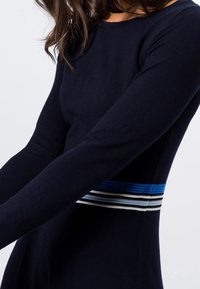 Marinblå långärmad klänning med figurnära silhuett, prydd med horisontella ränder i blått, grått och vitt vid midjan. Mjuk, slät textur.