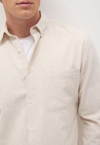 Camisa de botón color beige claro, con un collar relajado, bolsillo lateral y tejido texturizado. Lleva puesto una camiseta blanca debajo.