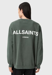 Oliwkowy sweter z długimi rękawami, z dużym białym logo "ALLSAINTS" oraz "LONDON" na plecach. Gładki, casualowy materiał.