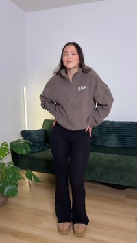 Pruun fliisist neljandiku luku ja logoga "GAP" dressipluus, mille all on mustad soonilised laienevad püksid ja beežid libisevad kingad. Taustal roheline diivan.