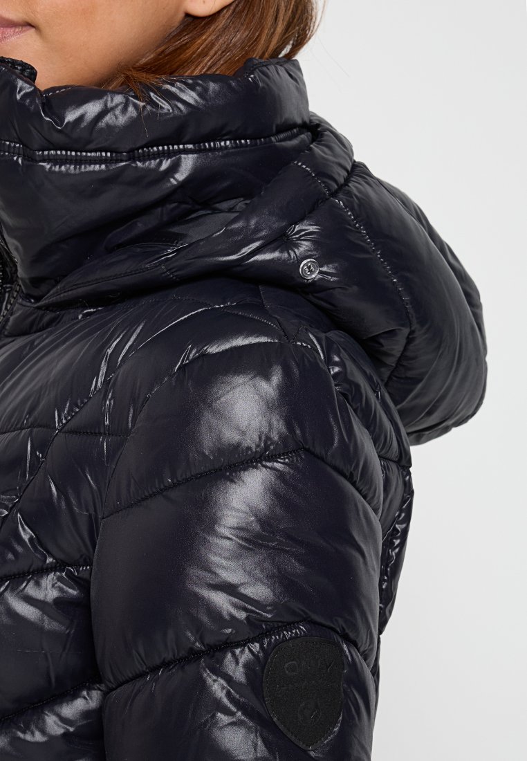 Zwarte pufferjack met een gequilte design, hoge kraag en een glanzende afwerking. Bevat een ronde zwarte logo-patch op de mouw.