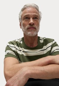 Zeleno-bílý pruhovaný tričko s krátkými rukávy. Světle pletě a šedé vlasy. Složené ruce vpředu, v uvolněné póze.