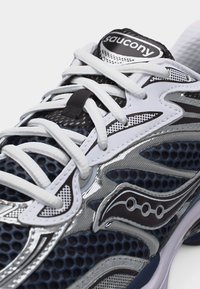 Μαύρο, άσπρο και ασημί παπούτσι τρεξίματος Saucony με επάνω μέρος από δίχτυ, άσπρα κορδόνια και λογότυπο της μάρκας στη γλώσσα και στην πλευρά.