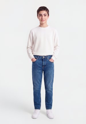 Adolescent debout les mains dans les poches, portant une chemise blanche à manches longues, un jean bleu et des baskets blanches sur un fond blanc uni.