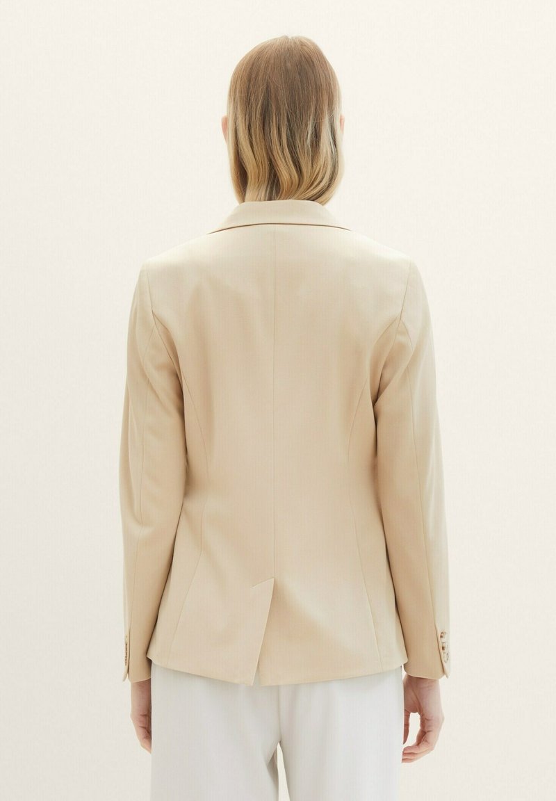 Blazer beige su misura con spalline, spacco singolo sul retro e polsini con bottoni. Tessuto liscio con una sottile trama.