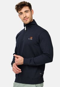 Sudadera de punto azul marino con cremallera de un cuarto, hecha de material suave, con un alto cuello, puños acanalados y un logo naranja en el pecho.
