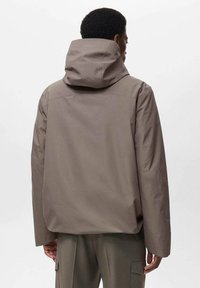Chaqueta marrón impermeable con capucha, con un ajuste relajado, textura suave y un diseño minimalista. Vista trasera que muestra las costuras de los hombros.